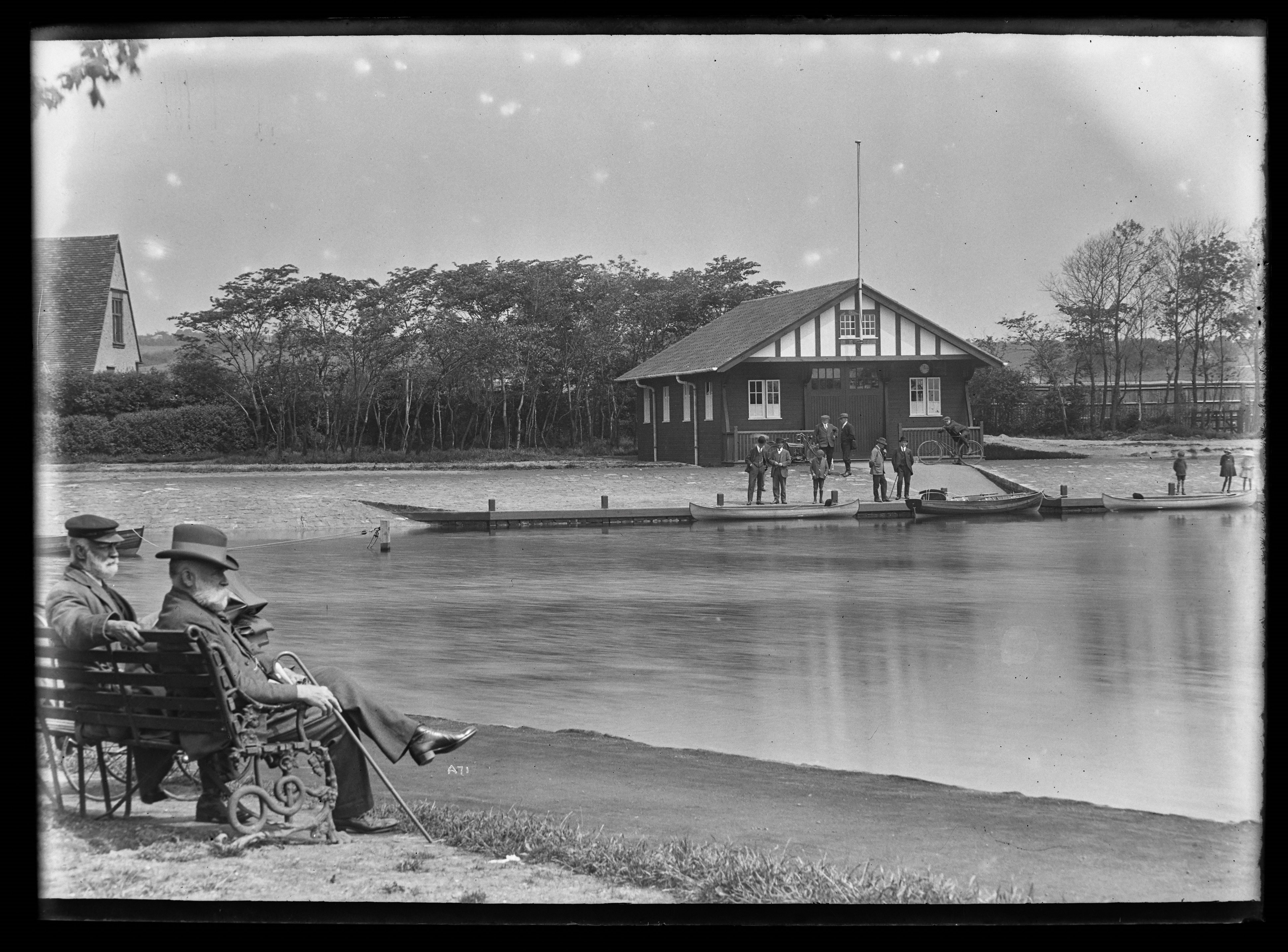 Black and white image of two men sat on a bench overlooking a boating lake